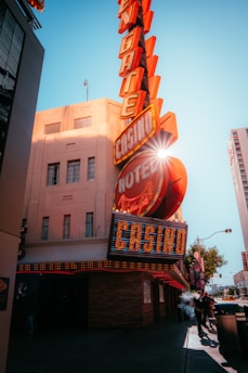 A vibrant city scene featuring a tall, illuminated casino sign with retro-style neon lights. The sign dominates the building facade with the words 'CASINO' and 'HOTEL'. Sunlight peeks through, creating a dramatic lens flare effect. The street is lively, with pedestrians casually walking and a hint of urban hustle.