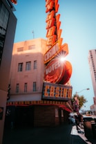 A vibrant city scene featuring a tall, illuminated casino sign with retro-style neon lights. The sign dominates the building facade with the words 'CASINO' and 'HOTEL'. Sunlight peeks through, creating a dramatic lens flare effect. The street is lively, with pedestrians casually walking and a hint of urban hustle.