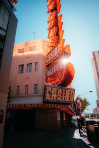 A vibrant city scene featuring a tall, illuminated casino sign with retro-style neon lights. The sign dominates the building facade with the words 'CASINO' and 'HOTEL'. Sunlight peeks through, creating a dramatic lens flare effect. The street is lively, with pedestrians casually walking and a hint of urban hustle.