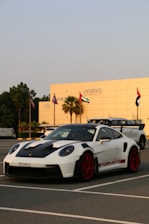 A white sports car with red rims and black accents is parked in a lot, with a modern building and several flags in the background. The car has a sleek design and the building reads 'Nawras Hospitality'.
