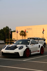 A white sports car with red rims and black accents is parked in a lot, with a modern building and several flags in the background. The car has a sleek design and the building reads 'Nawras Hospitality'.