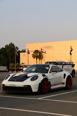 A white sports car with red rims and black accents is parked in a lot, with a modern building and several flags in the background. The car has a sleek design and the building reads 'Nawras Hospitality'.