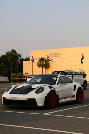 A white sports car with red rims and black accents is parked in a lot, with a modern building and several flags in the background. The car has a sleek design and the building reads 'Nawras Hospitality'.