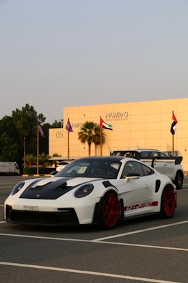 A white sports car with red rims and black accents is parked in a lot, with a modern building and several flags in the background. The car has a sleek design and the building reads 'Nawras Hospitality'.