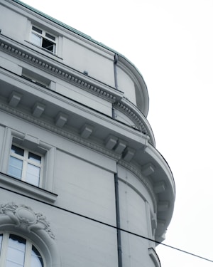A close-up view of an elegant building fa&ccedil;ade featuring architectural details such as decorative molding and a partially open window. The structure is built with smooth, light-colored stone and features arched windows with ornate designs.