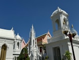A group of intricately designed white buildings with ornate architectural features stands against a deep blue sky. The structures display elements of religious or historical significance with spires and domes. Trees and well-maintained gardens surround the buildings, and a traditional lamp post with glass covers is visible in the foreground.
