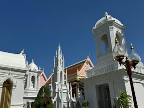 A group of intricately designed white buildings with ornate architectural features stands against a deep blue sky. The structures display elements of religious or historical significance with spires and domes. Trees and well-maintained gardens surround the buildings, and a traditional lamp post with glass covers is visible in the foreground.