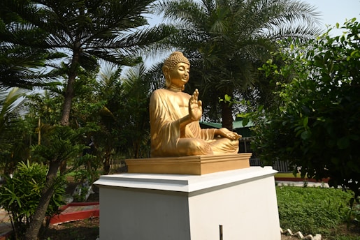 A golden statue of Buddha sitting in a meditative pose with one hand raised. It is placed on a white pedestal surrounded by lush green trees and foliage, under a clear sky.