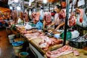A bustling meat market stall with various cuts of raw meat and sausages hanging and displayed on a wooden counter. Two vendors, one wearing an apron, are attending to the goods, surrounded by containers and bags filled with other produce. Bright lights illuminate the market scene, showcasing the fresh and raw meats prominently.