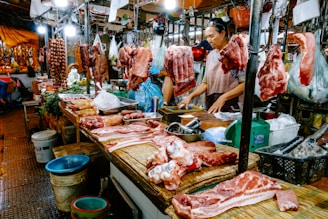 A bustling meat market stall with various cuts of raw meat and sausages hanging and displayed on a wooden counter. Two vendors, one wearing an apron, are attending to the goods, surrounded by containers and bags filled with other produce. Bright lights illuminate the market scene, showcasing the fresh and raw meats prominently.