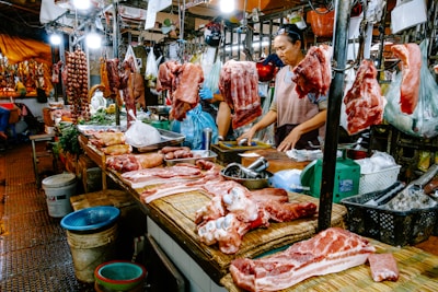 A bustling meat market stall with various cuts of raw meat and sausages hanging and displayed on a wooden counter. Two vendors, one wearing an apron, are attending to the goods, surrounded by containers and bags filled with other produce. Bright lights illuminate the market scene, showcasing the fresh and raw meats prominently.