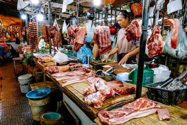 A bustling meat market stall with various cuts of raw meat and sausages hanging and displayed on a wooden counter. Two vendors, one wearing an apron, are attending to the goods, surrounded by containers and bags filled with other produce. Bright lights illuminate the market scene, showcasing the fresh and raw meats prominently.