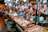A bustling meat market stall with various cuts of raw meat and sausages hanging and displayed on a wooden counter. Two vendors, one wearing an apron, are attending to the goods, surrounded by containers and bags filled with other produce. Bright lights illuminate the market scene, showcasing the fresh and raw meats prominently.