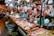A bustling meat market stall with various cuts of raw meat and sausages hanging and displayed on a wooden counter. Two vendors, one wearing an apron, are attending to the goods, surrounded by containers and bags filled with other produce. Bright lights illuminate the market scene, showcasing the fresh and raw meats prominently.