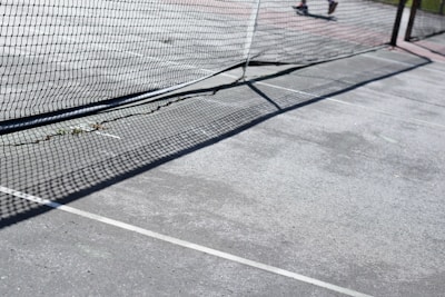 A tennis court with a net running across, casting intricate shadows on the concrete surface. The court lines are clearly visible, and part of a player's leg can be seen in the background.