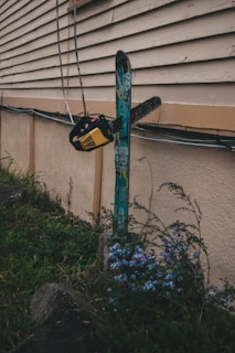 A chainsaw is embedded in a snowboard, which is fixed upright against an exterior wall of a building. The scene includes some overgrown grass and wildflowers in the foreground. The setting appears somewhat rugged and unconventional, with the equipment seeming out of context.