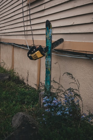 A chainsaw is embedded in a snowboard, which is fixed upright against an exterior wall of a building. The scene includes some overgrown grass and wildflowers in the foreground. The setting appears somewhat rugged and unconventional, with the equipment seeming out of context.