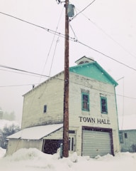 An old, weathered two-story building labeled 'Town Hall' stands amidst a snowy setting. The structure is wooden with peeling paint and a green trim on the roof. A utility pole with wires is prominently in front, and snow covers the ground deeply.
