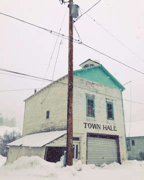 An old, weathered two-story building labeled 'Town Hall' stands amidst a snowy setting. The structure is wooden with peeling paint and a green trim on the roof. A utility pole with wires is prominently in front, and snow covers the ground deeply.