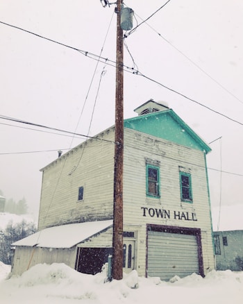 An old, weathered two-story building labeled 'Town Hall' stands amidst a snowy setting. The structure is wooden with peeling paint and a green trim on the roof. A utility pole with wires is prominently in front, and snow covers the ground deeply.
