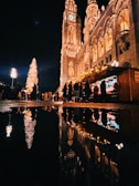 A festive night scene with a large, illuminated Christmas tree and a tall, ornate building lit up against the dark sky. A bustling crowd of people is gathered around holiday market stalls decorated with string lights. The reflections of the tree, building, and lights are visible in a puddle on the ground.
