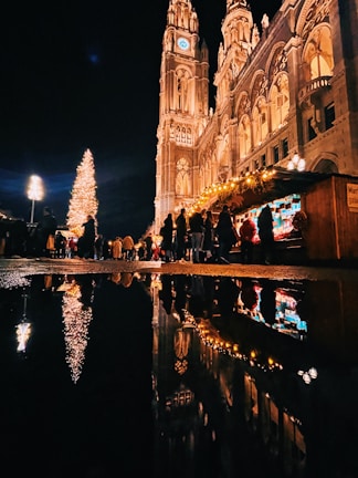 A festive night scene with a large, illuminated Christmas tree and a tall, ornate building lit up against the dark sky. A bustling crowd of people is gathered around holiday market stalls decorated with string lights. The reflections of the tree, building, and lights are visible in a puddle on the ground.