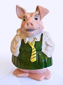 A ceramic figurine of a pig wearing a green dress and a yellow tie. The pig has distinctive features, including large ears and expressive eyes, giving it a whimsical and playful appearance.