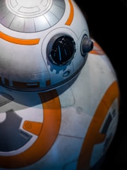 A close-up view of a spherical robot with a glossy black eye and intricate white and orange patterns. The mechanical design features circular elements and panels, suggesting a futuristic or sci-fi theme.