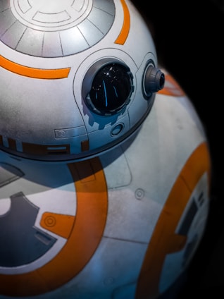 A close-up view of a spherical robot with a glossy black eye and intricate white and orange patterns. The mechanical design features circular elements and panels, suggesting a futuristic or sci-fi theme.
