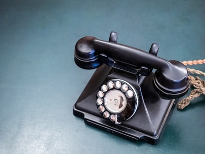 A vintage black rotary telephone with a coiled cord is placed on a flat, textured blue-green surface. The rotary dial is prominently featured, displaying numbers and letters.