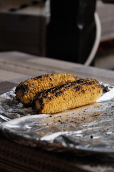 Two grilled corn cob pieces rest on a sheet of aluminum foil, showing charred marks and a golden-yellow color, placed on a wooden surface.