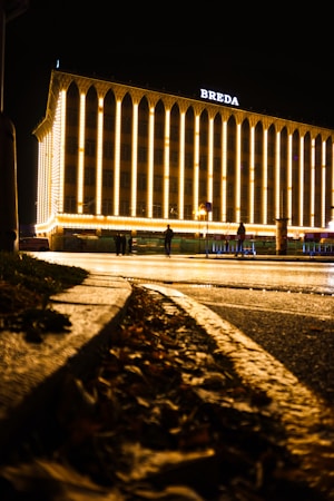 A building illuminated with warm, bright lights at night, displaying the word 'BREDA' on its top. The structure features prominent vertical columns with arches. In the foreground, fallen leaves cover a wet pavement, adding texture and depth to the scene.