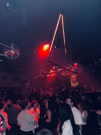 A crowded nightclub with people dancing and socializing under dim lighting. There are geometric light fixtures hanging from the ceiling and bright red lights add an intense ambiance. The room is filled with a lively crowd, with a mix of conversations and dance movements. In the background, there are discoballs and other decorative elements enhancing the vibrant atmosphere.