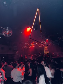 A crowded nightclub with people dancing and socializing under dim lighting. There are geometric light fixtures hanging from the ceiling and bright red lights add an intense ambiance. The room is filled with a lively crowd, with a mix of conversations and dance movements. In the background, there are discoballs and other decorative elements enhancing the vibrant atmosphere.