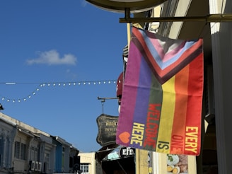 A diagonal flag with colorful stripes featuring the colors and design of the Progress Pride Flag, along with text that reads 'EVERYONE IS WELCOME HERE.' The flag is mounted on a building exterior along a street, and other buildings, signs, and string lights are visible in the background under a clear blue sky.