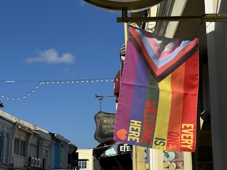 A diagonal flag with colorful stripes featuring the colors and design of the Progress Pride Flag, along with text that reads 'EVERYONE IS WELCOME HERE.' The flag is mounted on a building exterior along a street, and other buildings, signs, and string lights are visible in the background under a clear blue sky.
