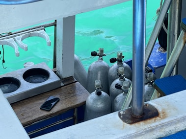 A collection of scuba diving tanks is arranged on a boat near a turquoise body of water. A mobile phone is placed on a wooden surface, and a set of clothes hangers are visible. The scene includes elements like metal bars and a platform with blue cushioning.