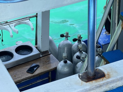 A collection of scuba diving tanks is arranged on a boat near a turquoise body of water. A mobile phone is placed on a wooden surface, and a set of clothes hangers are visible. The scene includes elements like metal bars and a platform with blue cushioning.
