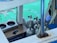 A collection of scuba diving tanks is arranged on a boat near a turquoise body of water. A mobile phone is placed on a wooden surface, and a set of clothes hangers are visible. The scene includes elements like metal bars and a platform with blue cushioning.