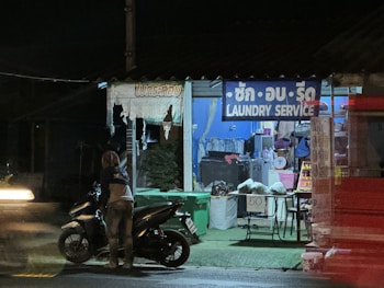 A small roadside laundry service is visible, with a sign in multiple languages. The shop is dimly lit, revealing washing machines inside. A person stands near a parked motorcycle on the street, which is illuminated by passing vehicle lights.