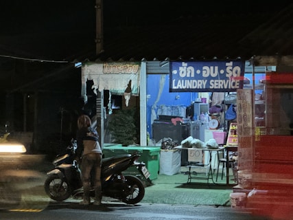 A small roadside laundry service is visible, with a sign in multiple languages. The shop is dimly lit, revealing washing machines inside. A person stands near a parked motorcycle on the street, which is illuminated by passing vehicle lights.