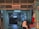 A dimly lit printing works area with a red sign that reads 'Sin & Lee Printing Works' in multiple languages. The entrance is an industrial corridor with fluorescent lighting and various industrial equipment scattered around. A person wearing a headscarf is walking past, captured with motion blur.