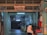 A dimly lit printing works area with a red sign that reads 'Sin & Lee Printing Works' in multiple languages. The entrance is an industrial corridor with fluorescent lighting and various industrial equipment scattered around. A person wearing a headscarf is walking past, captured with motion blur.