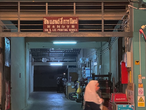 A dimly lit printing works area with a red sign that reads 'Sin & Lee Printing Works' in multiple languages. The entrance is an industrial corridor with fluorescent lighting and various industrial equipment scattered around. A person wearing a headscarf is walking past, captured with motion blur.