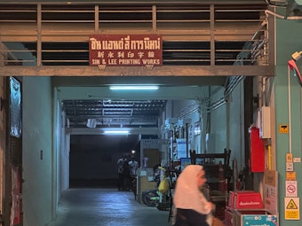 A dimly lit printing works area with a red sign that reads 'Sin & Lee Printing Works' in multiple languages. The entrance is an industrial corridor with fluorescent lighting and various industrial equipment scattered around. A person wearing a headscarf is walking past, captured with motion blur.