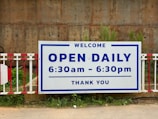 A large sign is prominently displayed with bold text indicating that the establishment is open daily from 6:30 am to 6:30 pm. The sign has a white background with blue border and text. It's mounted on a metal barrier and set against a rugged, brick-like background. Green foliage is visible at the bottom edge of the image.