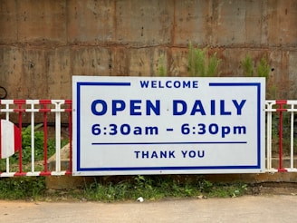 A large sign is prominently displayed with bold text indicating that the establishment is open daily from 6:30 am to 6:30 pm. The sign has a white background with blue border and text. It's mounted on a metal barrier and set against a rugged, brick-like background. Green foliage is visible at the bottom edge of the image.