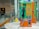 A brightly colored international payphone with orange, green, and yellow panels. It has a black handset attached with a metal cord and a numeric keypad with various instructions and buttons for operation. The phone is placed on a round marble surface with glass panels in the background displaying signs and reflections. The setting appears to be a public area, such as an airport or train station.
