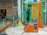 A brightly colored international payphone with orange, green, and yellow panels. It has a black handset attached with a metal cord and a numeric keypad with various instructions and buttons for operation. The phone is placed on a round marble surface with glass panels in the background displaying signs and reflections. The setting appears to be a public area, such as an airport or train station.