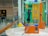 A brightly colored international payphone with orange, green, and yellow panels. It has a black handset attached with a metal cord and a numeric keypad with various instructions and buttons for operation. The phone is placed on a round marble surface with glass panels in the background displaying signs and reflections. The setting appears to be a public area, such as an airport or train station.